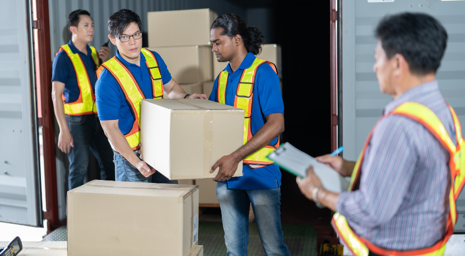 two warehouse workers unloading packages from a shipping container while someone checks paperwork
