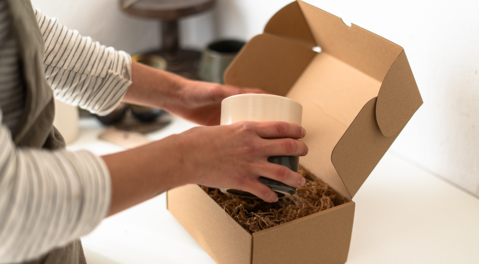 A pair of hands carefully lowering a ceramic pot into a cardboard box lined with cardboard/paper stuffing material
